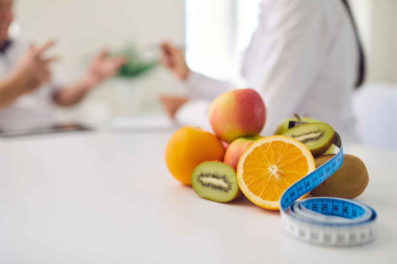 Fresh Fruit and Measuring Tape on Desk against Blurred Dietitian Giving Consultation to Patient